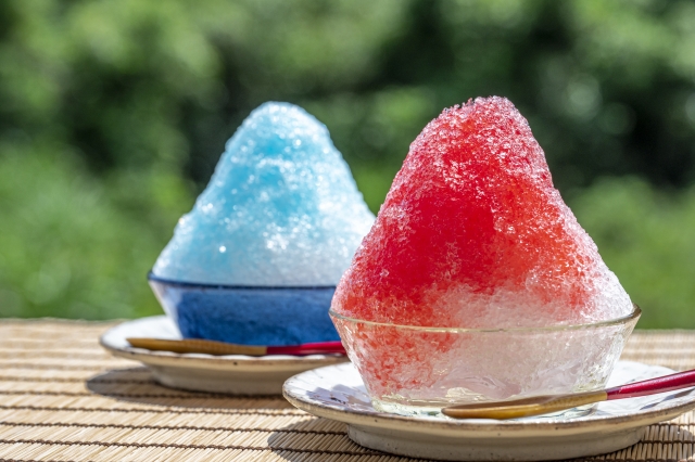 A photo of two glass bowls of kakigoori, a powdery ice dessert. One is topped with red syrup and the other is topped with blue syrup..