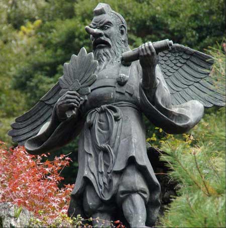 A photo of a tengu statue at a Shinto shrine in Japan. It looks like a humanoid goblin with a long nose, wispy beard, and a severe expression. It's brandishing a feathered fan in its right hand and has a pair of wings sticking out of its back.